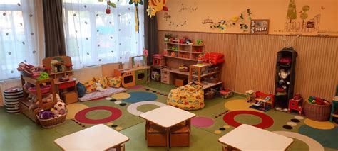 Egy bölcsődei csoportszoba, ahol gyerekek ülnek egy asztalnál és egy felnőtt mesét olvas nekik.