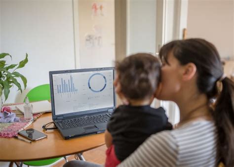 Egy dolgozó anya laptop előtt, mellette egy játszó gyermek, ami a távmunka és a rugalmasság lehetőségét jelképezi